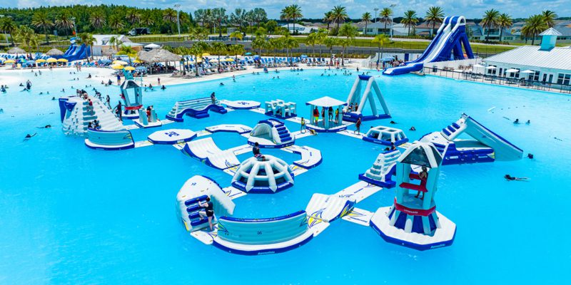 Inflatable obstacle course on top of clear blue water with climbing wall, bridges, slides, igloos, and various obstacles with kids in life vests playing. Beach in the background with guests lounging on beach chairs.