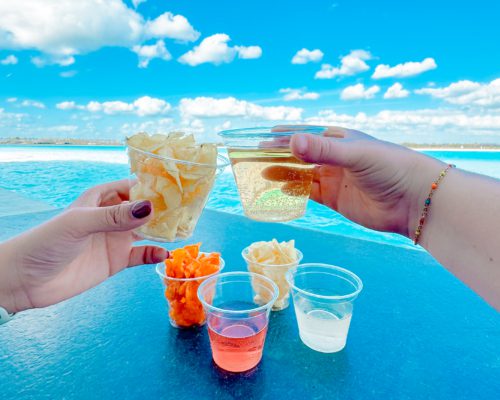 A cup of potato chips and a cup of white wine being held up against a blue sky with white clouds.